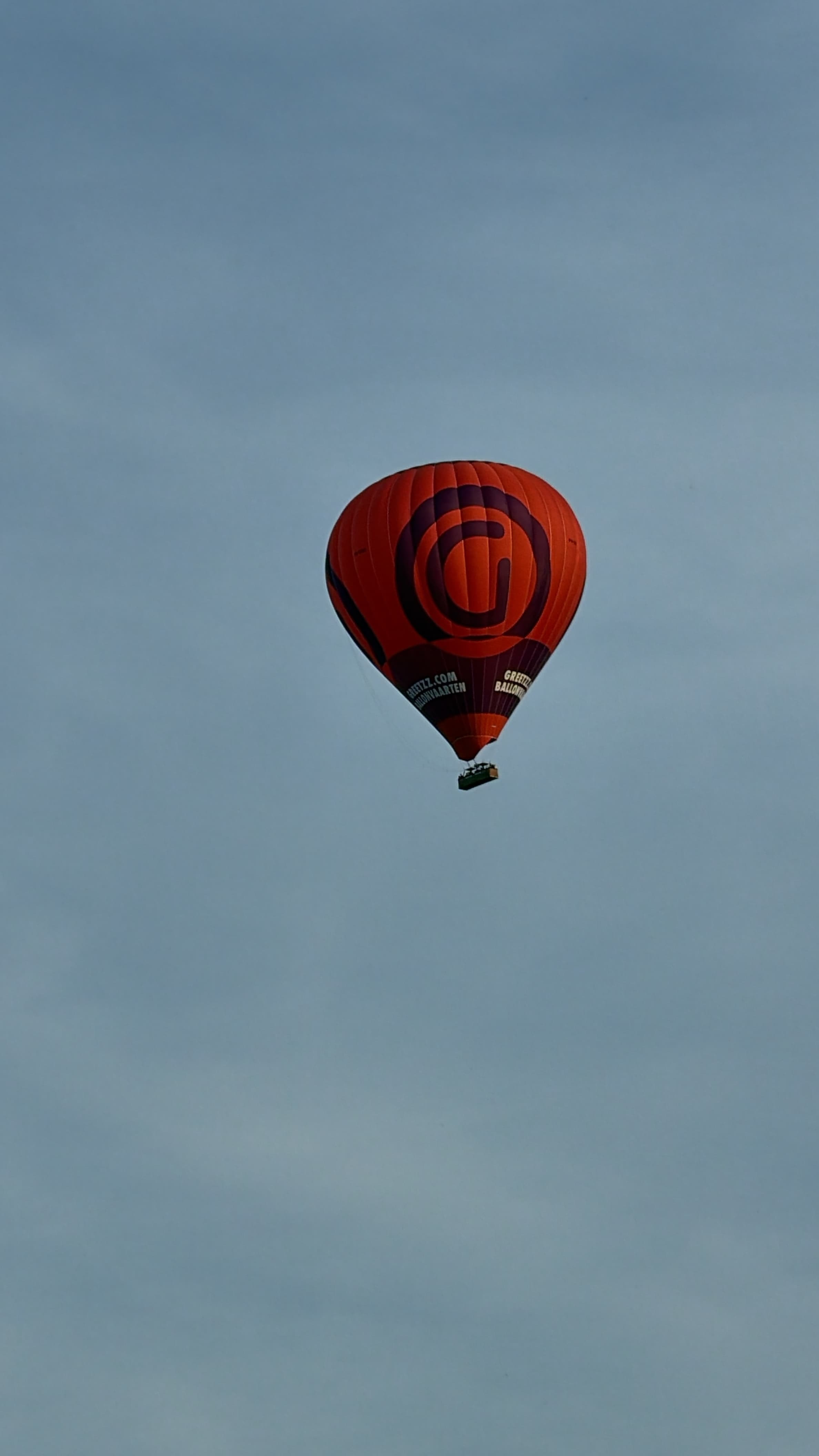 A red hot air balloon with a dark basket against a grey sky.