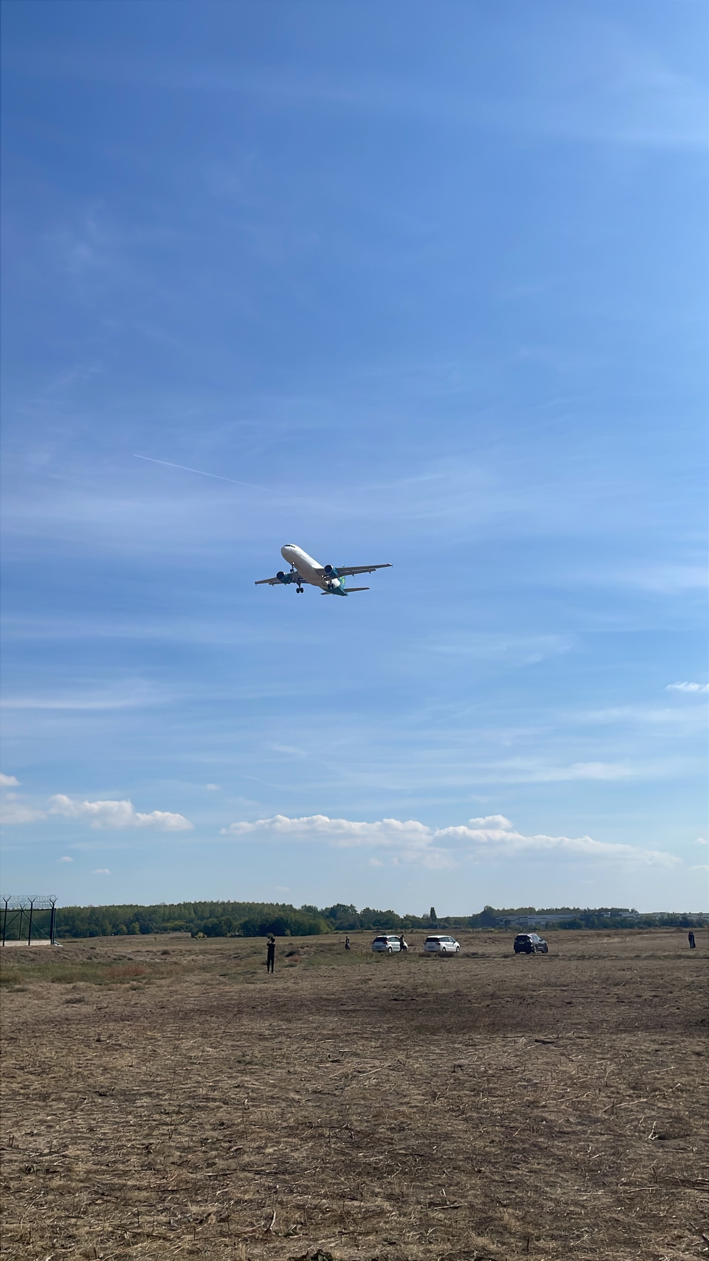 Airbus A321neo descending over open ground with parked cars below.